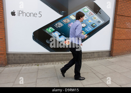 Ein Mann an Kopfhörer angeschlossen geht vorbei an einem großen Plakat für das iPhone 5 auf der Wand eines Einzelhändlers Carphone Warehouse. Stockfoto