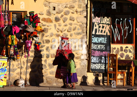 Einheimische indigene Dame in traditioneller Kleidung und Mädchen, die vorbei an Souvenirladen und Cafés geht, Ollantaytambo, Heiliges Tal, in der Nähe von Cusco, Peru Stockfoto