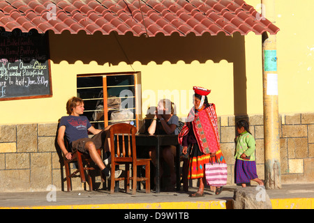 Einheimische Quechua-Ureinwohner in traditioneller Kleidung, die an Touristen vorbeiläuft, die im Straßencafé Ollantaytambo, Heiliges Tal, in der Nähe von Cusco, Peru sitzen Stockfoto