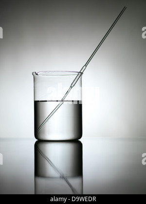 Becher gefüllt mit transparente Flüssigkeit mit eine Pipette, gegen einen Gradienten grauen Hintergrund und Verlegung auf einer reflektierenden Oberfläche Stockfoto