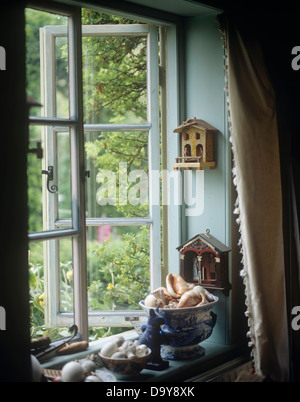 Nahaufnahme eines geöffneten Fensters mit Miniatur-Häuser an Wand über Porzellanschalen voller Muscheln am Schweller Stockfoto