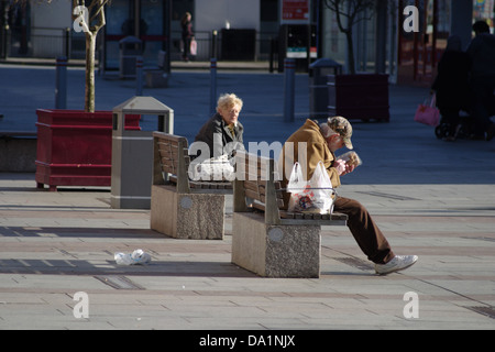 Mann und Frau sitzen auf Bänken im Stadtzentrum trennen. Man öffnet ein Paket von Lebensmitteln, hat eine Tragetasche. Frau hat Handtasche. Stockfoto