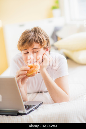 Teenager (14-15) auf Bett liegend, mit Laptop und Donut Essen Stockfoto