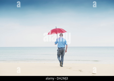 Mann geht auf einsamen Strand in Richtung Meer halten einen roten Regenschirm. Entfliehen Sie, Kontemplation, Zeitüberschreitung etc. Stockfoto