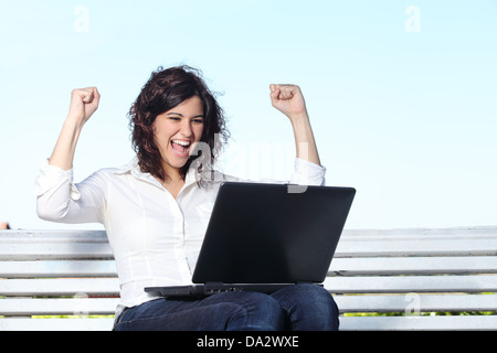 Euphorische Geschäftsfrau mit einem Laptop sitzen auf einer Bank mit dem Himmel im Hintergrund Stockfoto