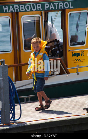 junger Bursche tragen Rettungsweste zu Fuß auf Ponton vorbei Hafen Patrouillenboot in Poole im Juni Stockfoto