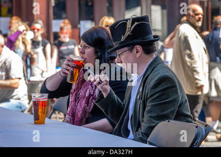 Mann mit einem Zylinder sitzt an einem Tisch vor einem Pub Überprüfung sein Telefon, während sein Partner einen Pint Bier trinkt, Stockfoto