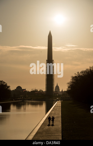 Washington D.C., Vereinigte Staaten – zwei Figuren mit Silhouetten gehen bei Sonnenaufgang am Rand des Lincoln Memorial Reflecting Pool entlang. Das Washington Monument und die Kuppel des US Capitols sind auf der Zentralachse der Mall ausgerichtet. Der McMillan-Plan von 1901–1902 legte diese Achse – vom Kapitol westwärts durch das Monument bis zu einem Terminaldenkmal an der Potomac-Küste – mit dem 2.028 Fuß (618 m) langen Pool als horizontaler Verbindungsglied fest, der auf Land gebaut wurde, das vor dem Ausbaggern des Army Corps in den 1870er Jahren als Wattenflächen diente Stockfoto