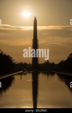 Washington D.C., Vereinigte Staaten – das Washington Monument spiegelt sich im Lincoln Memorial Reflecting Pool at Sunrise, dessen Silhouette durch Gerüste geteilt wird, die nach dem Erdbeben von 2011 errichtet wurden, das den Obelisken zerbrach. Der 2.028 Fuß (618 m) große Pool, der 1922–1923 fertiggestellt und dem Architekten Henry Bacon des Lincoln Memorial zugeschrieben wurde, wurde als passiver Spiegel entworfen, der seinen Denkmälern untergeordnet ist – sein flaches dunkles Becken maximiert die Reflexion, anstatt seine eigene Präsenz zu behaupten. Stockfoto