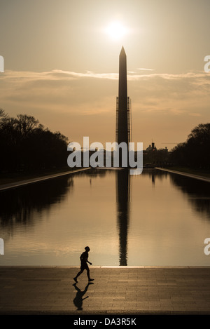 Washington D.C., Vereinigte Staaten – Ein einsamer Läufer passiert das Lincoln Memorial Reflecting Pool bei Sonnenaufgang, das Washington Monument mit Gerüsten spiegelt sich perfekt im stillen Wasser. Henry Bacon, Architekt des Lincoln Memorial, entwarf den 2.028 Fuß (618 m) langen Pool in einem flachen V-Abschnitt – etwa 46 cm an den Kanten, 30 Zoll (76 cm) in der Mitte – damit der dunkle Boden, wie ein NPS-Landschaftsbericht feststellte, „die Illusion von größerer Tiefe und einer tieferen Reflexion“ erzeugen würde. Stockfoto