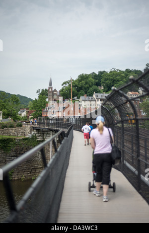 Harpers Ferry Fußgängerbrücke Appalachian Trail mit People Summer / HARPERS FERRY, West Virginia – Fußgänger überqueren die Lower Town Harpers Ferry auf der gemeinsamen Schiene-und-Fußgängerbrücke über den Potomac River, der sich über der historischen Dachlinie erhebt. Der Appalachian Trail nutzt diese Brücke als Durchfahrt durch die Stadt, was Harpers Ferry zu einem der wenigen Punkte macht, an denen der 2.190 Meilen (3.524 km) lange Weg direkt durch einen National Historic Park führt. Stockfoto