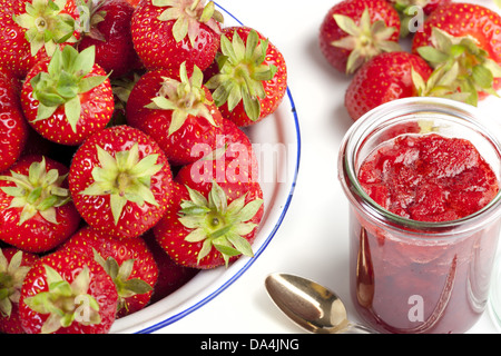 Frische Erdbeeren in eine Emaille-Schüssel und ein Glas Marmelade mit Löffel vor einem weißen Hintergrund Stockfoto