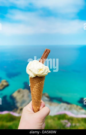 Hand, die Eiswaffel in Newquay, Cornwall, England, gegen das strahlend blaue Meer und den Himmel. Stockfoto