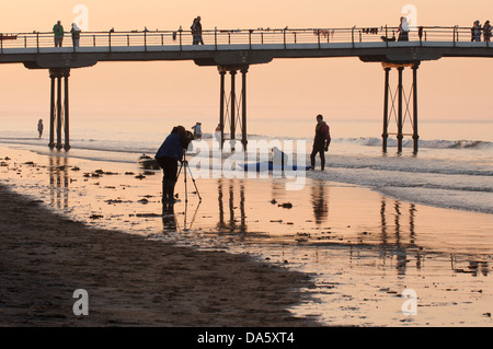 Farbenfrohe Sommer Sonnenuntergang Himmel & Sicht von Menschen zu Fuß am Meer Pier, Paddeln im Meer & Fotograf, Foto - Saltburn-by-the-Sea, England, UK. Stockfoto