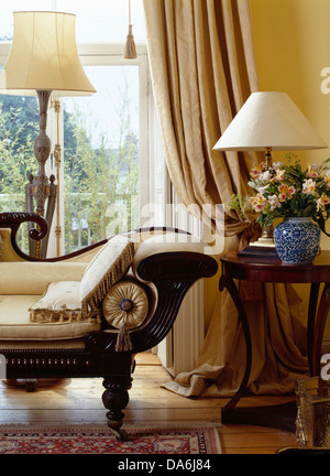 Vintage Chaiselongue und antiker Beistelltisch vor Fenster mit cremefarbenen Vorhängen im traditionellen Wohnzimmer Stockfoto