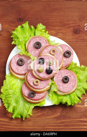 Brötchen mit Wurst, Salat und Oliven auf einer Platte auf Holztisch Stockfoto