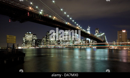 Brooklyn Bridge und die Skyline der Innenstadt Manhattan in der Dämmerung mit Lichtspuren von einem vorbeifahrenden Boot Stockfoto