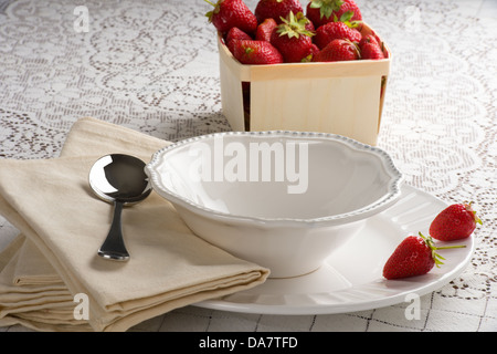 Leere Schüssel und Löffel auf Spitze Tischdecke warten auf Erdbeeren im Hintergrund im Karton Stockfoto