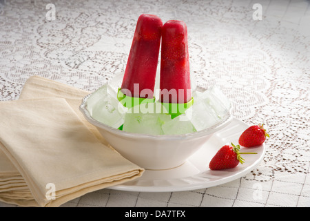 Hausgemachte Erdbeer Eis am Stiel sitzen in Schüssel mit Eis mit Karton Erdbeeren im Hintergrund und Servietten und Erdbeeren auf Platte Stockfoto