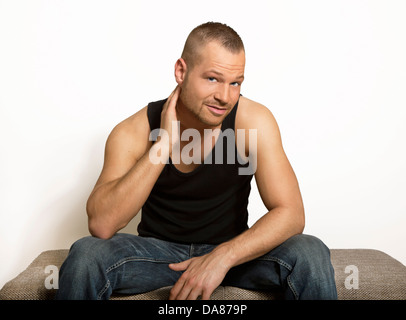 junger Mann sitzt mit einer Hand auf seinen Kopf, Blick auf die Kamera, Muskel-Shirt und Jeans, gut gepflegt und aufgebaut, Porträt Stockfoto