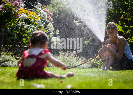 Ein Baby quietscht vor Freude, als sie von Mama sprühen Wasser aus einem Gartenschlauch abgekühlt ist. Stockfoto