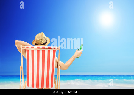 Mann mit Hut sitzt auf einem Liegestuhl und hält eine Bierflasche am Strand Stockfoto