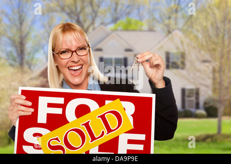 Glückliche junge Frau mit verkauft für Immobilien Verkaufsschild und Schlüssel vor Haus. Stockfoto