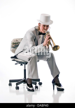 Hübsche blonde Frau in Smoking und Zylinder spielen einer Trompete auf einem Hocker sitzend. Stockfoto