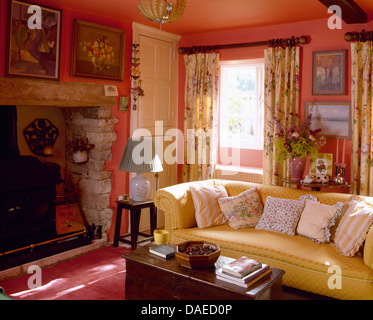 Gelbes Sofa mit floralen Kissen in fröhlichen roten Land Wohnzimmer mit Kamin Inglenook und floral Gardinen Stockfoto