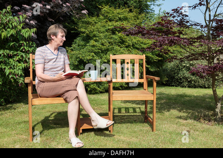 Ältere Frau, die Witwe von Baby boomer Generation traurig und einsam allein neben einem leeren Begleiter Sitz in sonniger Garten im Sommer sitzen. Großbritannien Stockfoto