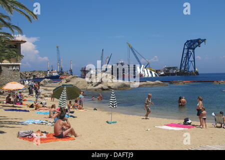 Giglio, Italien. 10. Juli 2013. Menschen genießen Sie die Sonne am Strand mit dem Wrack des Kreuzfahrtschiffes "Costa Concordia" im Hintergrund auf der Insel Giglio, Italien, 10. Juli 2013. Foto: MIRIAM SCHMIDT/Dpa/Alamy Live News Stockfoto