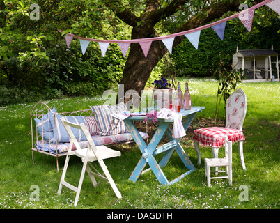 Hübsche Girlanden oben blau lackierten Tisch zum Mittagessen unter Baum auf Rasen Vintage Tagesbett gestapelt mit Kissen Stockfoto