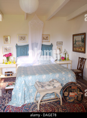 Moskitonetz über dem Bett mit gemalten blauen Kopfteil und blaue + weiße Abdeckung in französischer Landhaus Schlafzimmer mit Werkzeug und Hut Whitebox Stockfoto