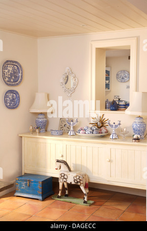 Blau + weiß Keramik auf weiß lackierte Sideboard unter Luke im Land Esszimmer mit alten blauen Holzkiste und Holzpferd Stockfoto