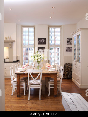 Weiße Stühle bei einfachen Holztisch in modernen weißen Küche Esszimmer mit Parkettboden und weiße Plantage Fensterläden Stockfoto