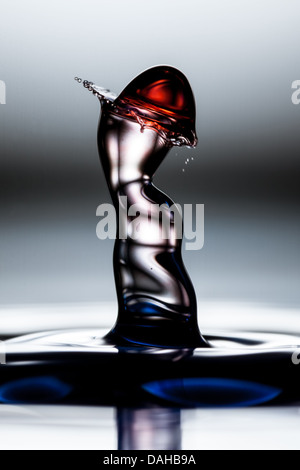 Ein Hochgeschwindigkeitsfoto nimmt einen einzelnen Tropfen dunkler Flüssigkeit auf, der auf eine Oberfläche trifft und einen hohen, skulpturalen Spritzer erzeugt. Der Spritzer bildet eine abstrakte, kurvenförmige Form, die einer Figur ähnelt, mit einem Stockfoto