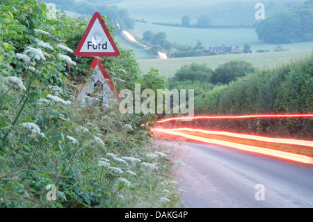 Englische Landschaft. Die roten Rückleuchten eines Auto-Streifen durch die grüne Hecken einen schmalen Feldweg in Dorset. Ein Schild warnt vor einer Furt. Stockfoto