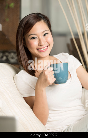 Schöne chinesische Frau hält eine Kaffeetasse beim Sitzen auf ihrem sofa Stockfoto