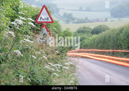 Englische Landschaft. Die roten Rückleuchten eines Auto-Streifen durch die grüne Hecken einen schmalen Feldweg in Dorset. Ein Schild warnt vor einer Furt. Stockfoto