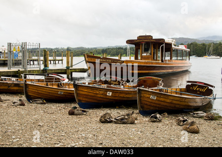 Einige Ruderboote und ein Fahrgastschiff am Ufer des Lake Windermere neben hölzerne Pier. Graue Wolken überhängenden entfernten Hügel. Stockfoto