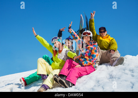 Glückliche Freunde Männer und Frauen sitzen im Schnee mit Snowboards heben und winken Hände Stockfoto