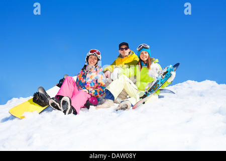 Drei glückliche Freunde Mann und Frauen sitzen im Schnee mit Snowboards heben und winken Hände Stockfoto