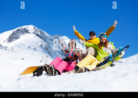 Drei glückliche Freunde Mann und Frauen sitzen im Schnee mit Snowboards heben und winken Hände Stockfoto