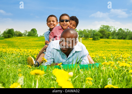 Glücklich lächelnd schwarze Afrikaner mit jungen sitzt auf seinem Rücken legen im Frühjahr Park Bereich mit gelben Löwenzahn Stockfoto