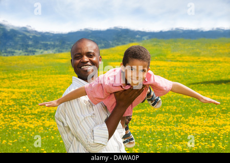 Schwarze Vater spielen mit kleinen Jungen hält ihn im Flugzeug Pose im Frühjahr gelbe Löwenzahn Feld Stockfoto