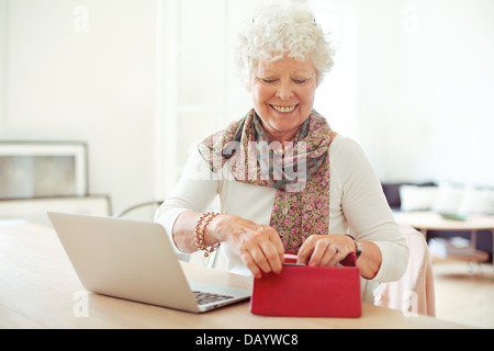 Fröhliche alte Frau vor Laptop immer etwas aus ihrer Brieftasche Stockfoto