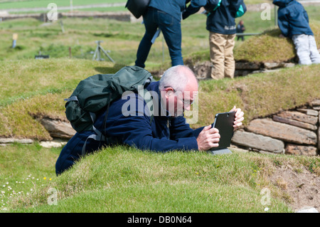 Ein Tourist, der einen Tablet-Computer als Digitalkamera verwendet, um ein Foto bei Skara Brae zu machen. Senior mit moderner Technologie.