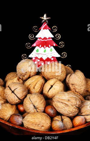 Holzschale voller Walnüsse und Haselnüsse mit einem ziemlich Zinn Weihnachtsbaum nach hinten gegen einen schwarzen Hintergrund. Stockfoto