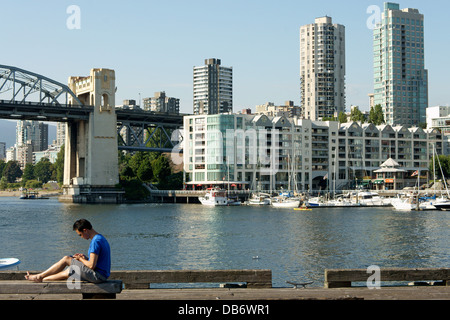 Junge asiatische Mann Überprüfung seiner Smartphone beim Sitzen auf einem Pier, Granville Island, Vancouver, Britisch-Kolumbien, Kanada Stockfoto
