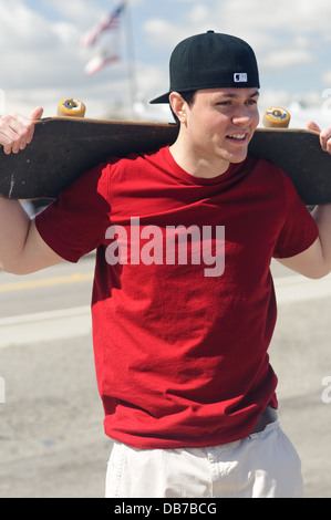 Gemischtrassig junger Mann hält Skateboard über Schulter lächelnd outdoor Portrait blauen Himmelshintergrund Stockfoto